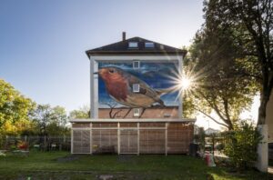 Das Wandbild von Denis Klatt zeigt ein überdimensionales Rotkehlchen in einer surrealen Landschaft aus Wüste und Meer - realistisch inszeniert mit lebendiger Licht- und Schattensetzung. (Foto: LWL / Maxime P. Lindenbaum)