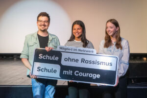 Die Schülerinnen Aurelie (Mitte) und Clara aus der 10. Jahrgangsstufe nahmen das Schild aus den Händen von Oliver Kottmann vom Netzwerk „Schule ohne Rassismus – Schule mit Courage“ entgegen. (Foto: Bischöfliche Pressestelle/Ann-Christin Ladermann)