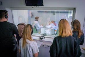 Teilnehmende des Trainings beobachten die Visite, die Medizinstudierender Mark Schlarmann durchführt, aus dem Nebenraum mit einer semitransparenten Scheibe. (Foto: UKM / Wibberg)
